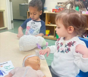 Two toddlers playing with dolls and a stuffed animal indoors.