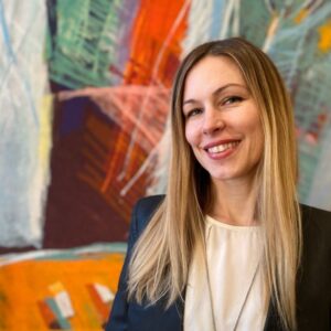 Smiling woman with long blonde hair in front of colorful abstract art.
