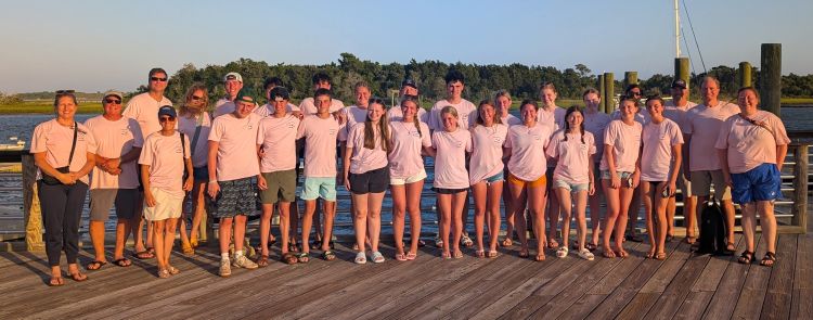 A group of people standing on a wooden dock at sunset wearing light pink shirts.