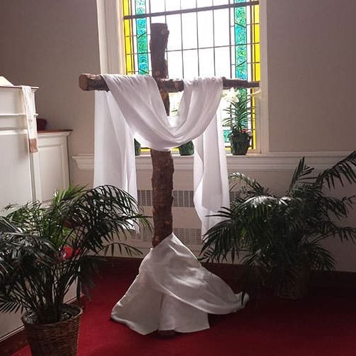 A white cloth draped over a wooden cross in front of a stained glass window.