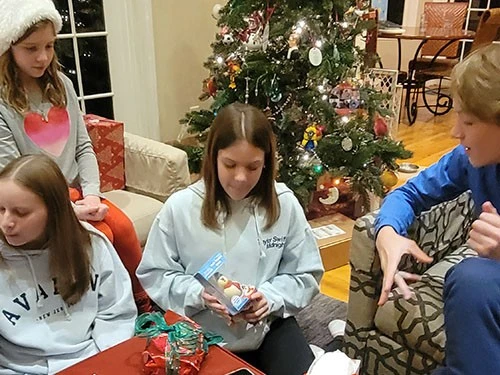 A girl opening a gift by the Christmas tree with family around.