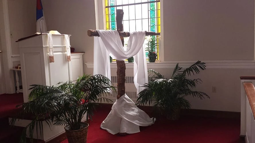 A white cloth draped over a wooden cross in front of a stained glass window.