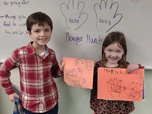 Two children hold orange papers with handprints in a classroom.