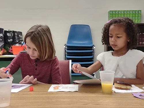 Two children painting at a table with art supplies and juice cups.