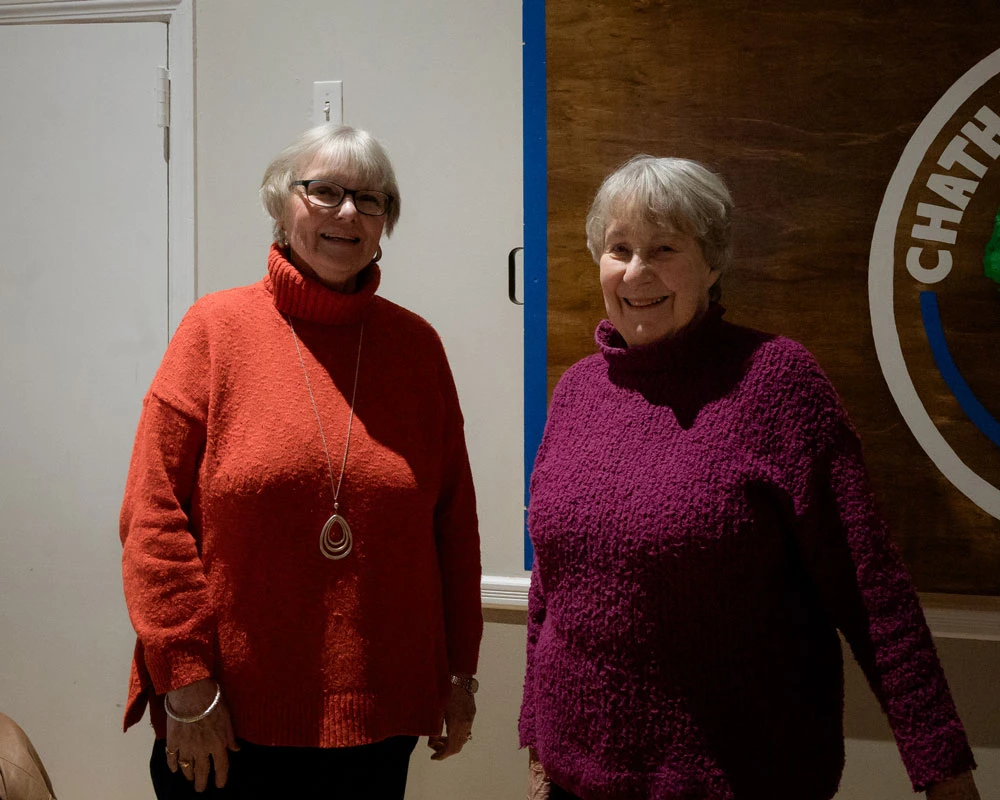 Two elderly women smiling, wearing cozy sweaters indoors.
