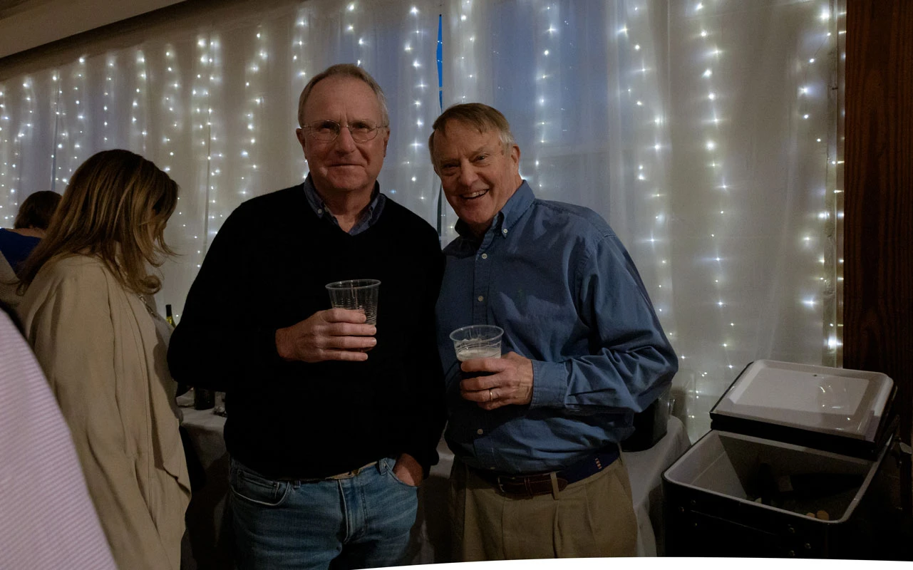 Two men smiling, each holding a drink in a warmly lit setting with string lights.