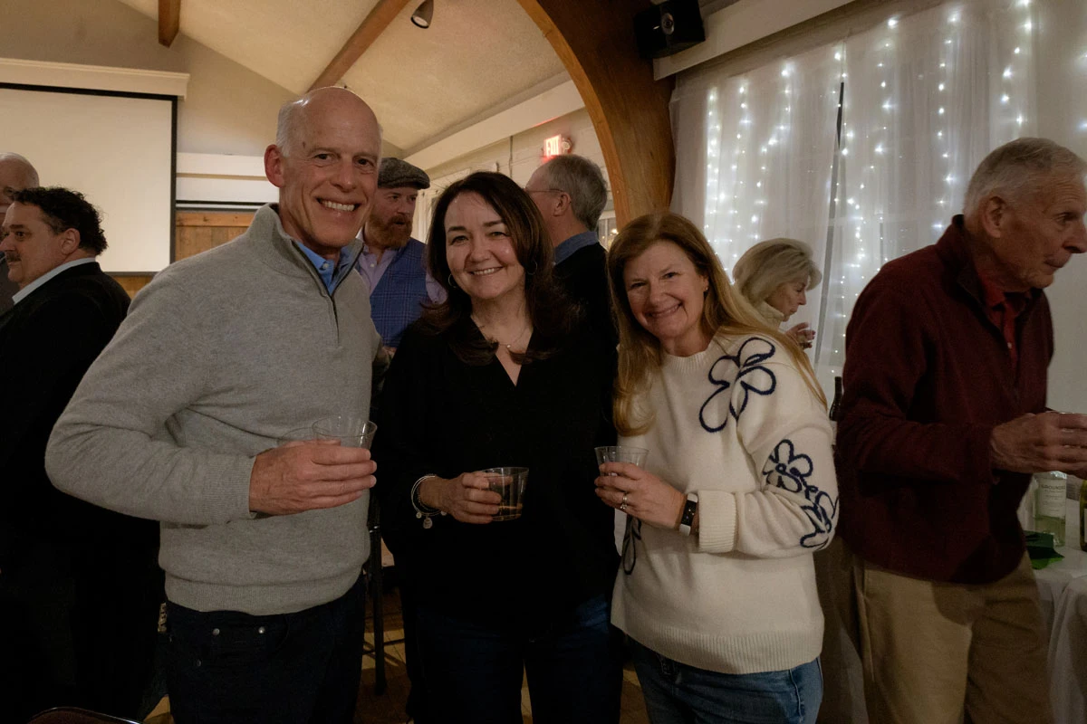 Three friends smiling and holding drinks at a cozy indoor gathering.
