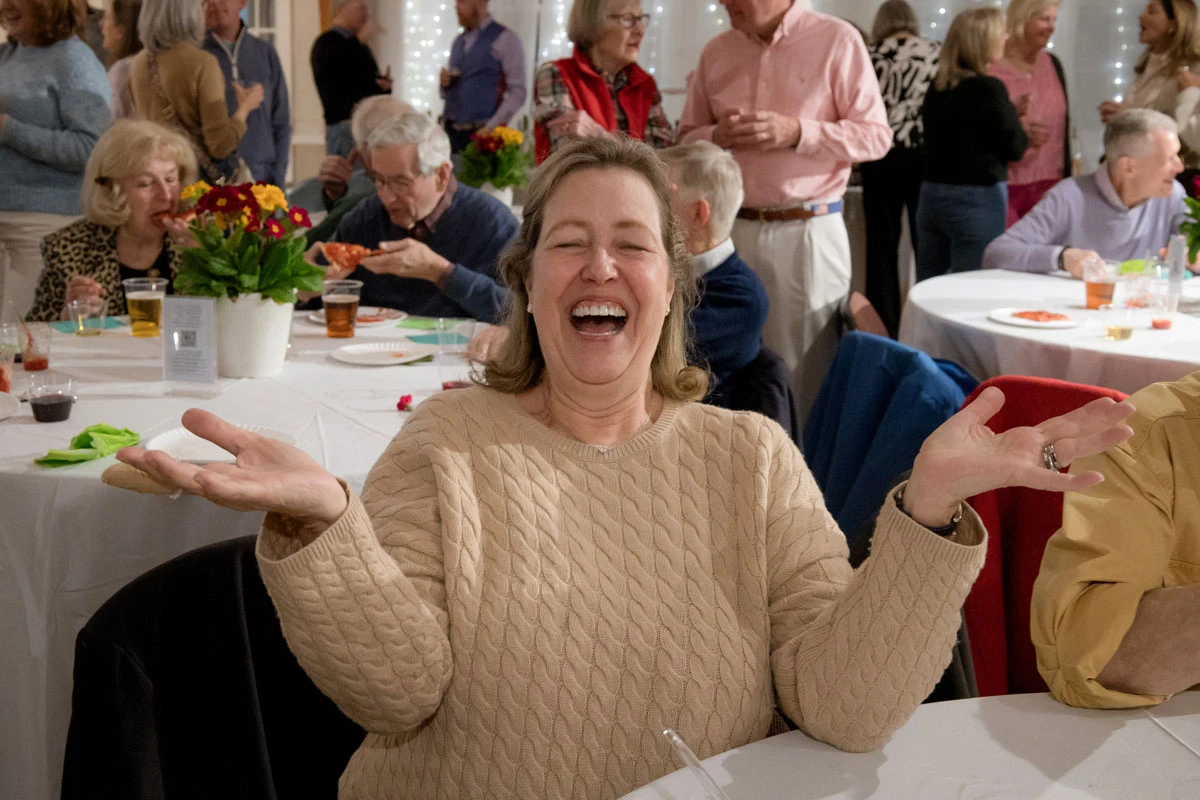 A joyful woman laughing with arms raised at a social gathering.