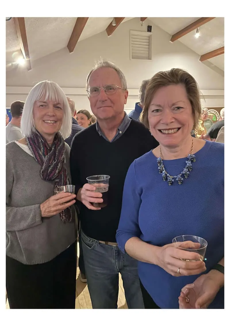 Three adults smiling and holding drinks indoors.
