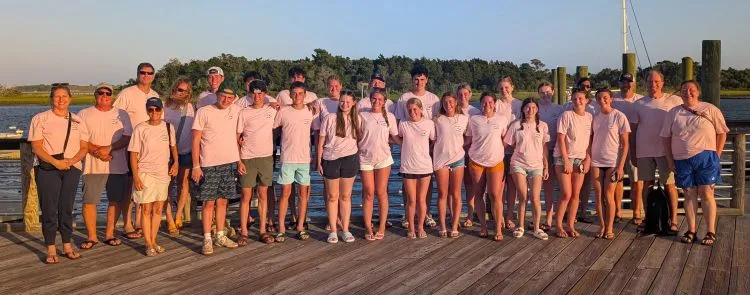 A group of people standing on a wooden dock at sunset wearing light pink shirts.