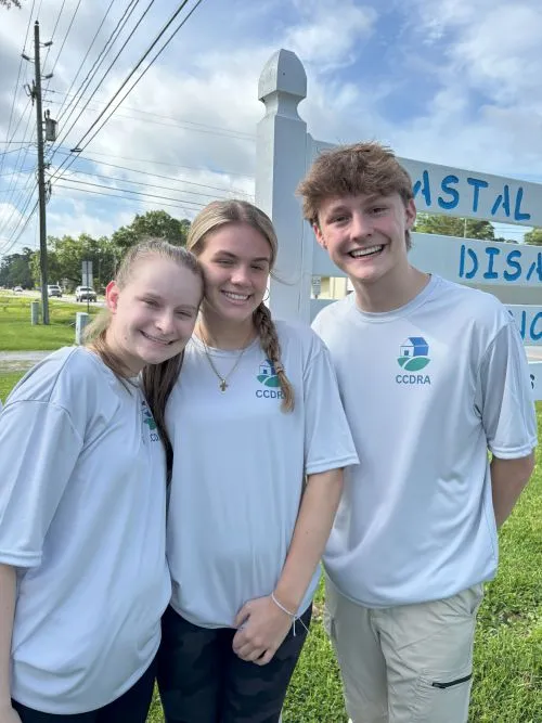 Three smiling teenagers in white shirts posing outdoors on a sunny day.