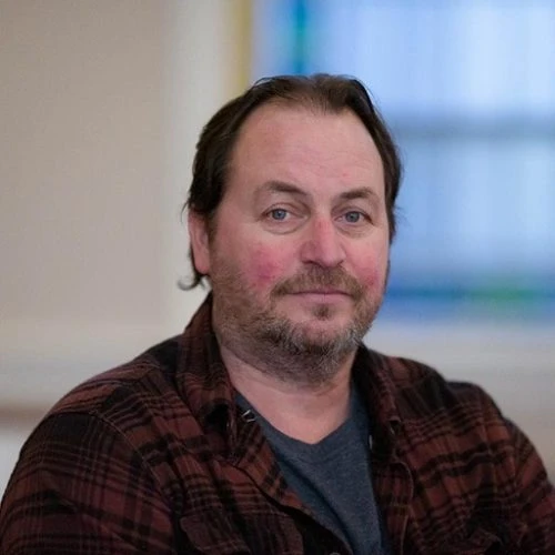 Portrait of a man with medium-length hair and a beard, wearing a plaid shirt.