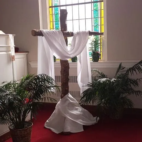 A white cloth draped over a wooden cross in front of a stained glass window.