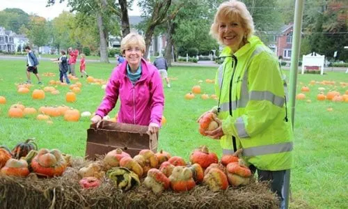 Two women arranging pumpkins on hay bales at an outdoor autumn event.